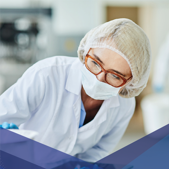 Laboratory technician wearing a mask, hair cover, and glasses, working in a clinical or sterile environment.