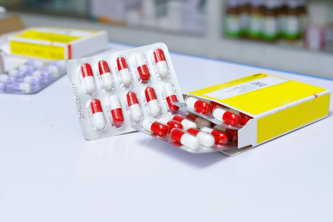 Plaquettes thermoformées de gélules rouges et blanches partiellement sorties d’une boîte avec une étiquette jaune.