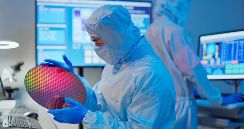 Técnico de semicondutores usando equipamento de proteção completo para sala limpa e luvas azuis inspecionando um wafer de silício refletivo, com telas de monitoramento digital e outro técnico trabalhando em segundo plano.